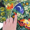 Na zdjęciu widać fragment gry planszowej z kolorowymi ilustracjami przedstawiającymi rośliny, kwiaty i księżyc na niebieskim tle. W centrum znajduje się dłoń z pomalowanym na czerwono paznokciem, wskazująca na jeden z elementów planszy.