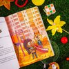 Na zdjęciu widać otwartą książkę z ilustracją przedstawiającą plac zabaw z postaciami zwierząt na tle wysokiego budynku. Wokół książki znajdują się różne przedmioty, takie jak żółty wiatraczek, kostka do układania, lupa, pluszowa zabawka, piłka i sztuczne