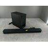 Czarny soundbar i subwoofer ustawione na szarej powierzchni, z widocznymi kablami zasilającymi. Na soundbarze znajduje się żółta strzałka skierowana w stronę subwoofera.
