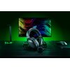 Na zdjęciu widoczne są czarne słuchawki nauszne z mikrofonem, umieszczone na biurku przed monitorem z kolorowym, abstrakcyjnym tłem i logo Razer. Obok znajdują się myszka, podkładka oraz stojak na słuchawki, wszystko w ciemnej kolorystyce z zielonymi akce