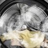 Widoczny jest bęben pralki z wnętrzem wypełnionym parą i kolorowymi tkaninami. Wnętrze bębna ma metaliczną, perforowaną powierzchnię.