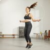 Na zdjęciu znajduje się osoba skacząca na skakance w jasnym pomieszczeniu z metalową szafką, niskim stolikiem i rośliną w tle. Osoba ubrana jest w sportowy strój, a jej włosy unoszą się w powietrzu.