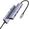 Na zdjęciu widoczny jest hub USB typu C w kolorze srebrnym z kilkoma portami, w tym HDMI i USB, oraz kablem zakończonym wtykiem USB-C. Na urządzeniu znajduje się napis "UGREEN 4K 60HZ".