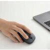 Na zdjęciu widać dłoń trzymającą czarną, bezprzewodową mysz komputerową na białym biurku obok szarego laptopa. Klawiatura laptopa jest częściowo widoczna, a mysz ma prosty, ergonomiczny kształt.