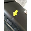 Czarny grill z widocznymi otworami wentylacyjnymi i żółtą strzałką wskazującą na wgniecenie na powierzchni. Na górze znajduje się metalowy element przypominający pokrętło.