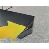 Na zdjęciu widoczny jest czarny soundbar z perforowaną powierzchnią, na który wskazuje żółta strzałka. Tło stanowi szara powierzchnia przypominająca dywan.