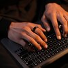 Na zdjęciu widoczne są dłonie osoby piszącej na podświetlonej klawiaturze laptopa. Na jednym z palców znajduje się ciemny pierścień.