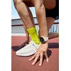 Osoba w sportowym stroju przygotowuje się do biegu na bieżni, nosząc biały smartwatch na nadgarstku i białe buty sportowe. Na skarpetkach widoczny jest czarny napis "UA".