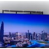 Na zdjęciu widoczny jest telewizor wyświetlający panoramę miasta z wysokimi wieżowcami i błękitnym niebem. W dolnej części ekranu znajduje się obszar z wodą otoczony budynkami.