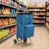 Niebieski wózek na zakupy z dużą komorą i kieszenią z przodu stoi na podłodze w sklepie spożywczym, otoczony półkami z kolorowymi opakowaniami produktów. W tle widoczne są rzędy półek z różnorodnymi artykułami spożywczymi.
