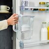 Dzbanek filtrujący z białym uchwytem jest wkładany do lodówki, na której drzwiach znajdują się różne produkty, w tym butelki z mlekiem i sokiem oraz słoiki z przetworami. W tle widoczna jest półka z kubkiem.