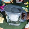 Na zdjęciu znajduje się dzbanek filtrujący z szarym wieczkiem, na którym widnieje logo "BRITA". W tle widać różne przedmioty, takie jak kolorowe koraliki, kubek z przyborami i talerzyk.