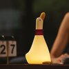 Na zdjęciu znajduje się lampka nocna w kształcie kaczki, świecąca ciepłym, żółtym światłem, z czerwonym elementem na szyi. Obok lampki widoczny jest numer "27" na stojaku.