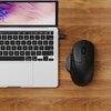 Na zdjęciu widać czarną mysz komputerową obok otwartego laptopa na drewnianym blacie. Ekran laptopa wyświetla abstrakcyjny wzór w odcieniach fioletu i różu, a klawiatura ma czarne klawisze z białymi oznaczeniami.