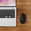 Na zdjęciu widoczny jest fragment otwartego laptopa z klawiaturą i touchpadem oraz czarna mysz komputerowa na drewnianym blacie. Ekran laptopa wyświetla abstrakcyjny wzór w odcieniach różu i fioletu.