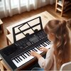 Na zdjęciu widoczny jest czarny keyboard z nutami na pulpicie, na którym gra dziecko. Instrument ma wyświetlacz i liczne przyciski funkcyjne.