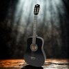 Czarna gitara klasyczna stoi na drewnianej powierzchni, oświetlona z góry, z widocznym logo "V-TONE" na główce. W tle znajduje się rozmyte, ciemne tło z efektem świetlnym.