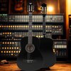Czarna gitara klasyczna z widocznym logo "V-TONE" na główce, ustawiona na tle panelu z licznymi pokrętłami i kontrolkami. Obok niej znajduje się druga gitara, pokazana od tyłu, na drewnianej podłodze.
