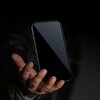Na zdjęciu widoczna jest dłoń trzymająca smartfon z ciemnym ekranem, na czarnym tle. Ekran telefonu jest wyłączony lub w trybie uśpienia, nie pokazując żadnych treści.