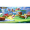Na obrazku widoczna jest kolorowa scena z gry "Mario + Rabbids Kingdom Battle" z postaciami w dynamicznych pozach na tle zielonego krajobrazu z ceglastymi blokami i niebieskim niebem. W lewym górnym rogu znajduje się logo gry z napisem "Mario + Rabbids Ki