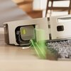 Biały robot sprzątający z widocznym mechanizmem czyszczącym i zielonym światłem skierowanym na podłogę, obok dywanu. Na urządzeniu znajduje się napis "PerfectEdge".
