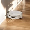 Na zdjęciu widoczny jest biały robot sprzątający na drewnianej podłodze, obok którego leżą rozsypane okrągłe płatki. Robot znajduje się przy ścianie, a na podłodze widać cienie.