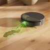 Na zdjęciu znajduje się czarny robot sprzątający na drewnianej podłodze, emitujący zielone światło na zabrudzenie przed nim. Światło tworzy siatkę na powierzchni podłogi, wskazując obszar do czyszczenia.