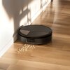 Czarny robot sprzątający znajduje się na drewnianej podłodze obok ściany, zbierając rozsypane okrągłe płatki. Na podłodze widać cienie, które tworzą wzory.