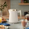 Na zdjęciu znajduje się czajnik o żebrowanej powierzchni z metalicznymi elementami, na którym widnieje napis "Russell Hobbs". W tle widać drewniane półki z dekoracjami, a na blacie znajdują się szklanka soku, filiżanka z napojem oraz talerz z pieczywem i 