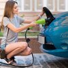 Na zdjęciu widoczna jest osoba podłączająca zieloną ładowarkę do samochodu elektrycznego, której wtyczka jest umieszczona w gnieździe ładowania niebieskiego samochodu. Na ładowarce znajduje się napis "Qoltec".