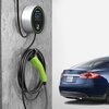Na zdjęciu widoczna jest ładowarka do samochodu elektrycznego zamontowana na ścianie, z zielonym uchwytem i zwiniętym kablem. Obok znajduje się niebieski samochód z tablicą rejestracyjną "MODEL S".
