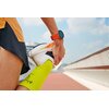 Osoba w sportowym stroju rozciąga nogę, trzymając stopę w ręce, na której znajduje się pomarańczowy smartwatch. W tle widoczny jest fragment bieżni oraz niebo z chmurami.