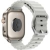 Srebrny smartwatch z okrągłym przyciskiem z boku, posiadający jasny, falisty pasek z metalową klamrą. Na spodzie zegarka widoczne są czujniki oraz dwa małe otwory.