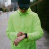 Osoba w jaskrawozielonej kurtce i czapce z daszkiem patrzy na czarny smartwatch na nadgarstku. W tle widoczna jest rozmyta ulica z budynkami i roślinnością.