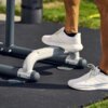 Na zdjęciu widoczna jest część zewnętrznej stacji treningowej z metalowymi elementami, na której stoi osoba w białych butach sportowych. Tło stanowi zielona trawa i czarna mata treningowa.