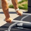 Na zdjęciu widoczne są ręce trzymające metalowe uchwyty zewnętrznej stacji treningowej na tle betonowej nawierzchni i zieleni. Na nadgarstku znajduje się biały zegarek sportowy.