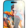 Zdjęcie przedstawia smartfon z nałożonym szkłem hartowanym, które redukuje refleksy świetlne, a na ekranie widoczna jest kolorowa fotografia osoby w okularach przeciwsłonecznych. W tle widać jasne niebo i słońce, które podkreślają właściwości antyrefleksy