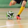 Na zdjęciu widać piłkę nożną z zielono-białym wzorem na hali sportowej oraz nogi osoby w żółtych getrach i różowych butach sportowych, kopiącej piłkę. Podłoga jest jasna, a w tle widoczna jest rozmyta zielona ściana.