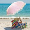 Na zdjęciu widać czerwony parasol ogrodowy z frędzlami, rozłożony na piaszczystej plaży przy brzegu morza. Pod parasolem znajduje się osoba siedząca na leżaku, przykryta kolorowym ręcznikiem.