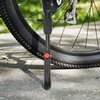 Na zdjęciu widoczna jest stopka rowerowa zamontowana przy tylnym kole roweru, stojąca na betonowej powierzchni. Stopka ma czarny kolor z czerwonym elementem regulacyjnym, a w tle widać fragmenty roweru i rozmytą zieleń.