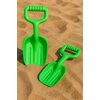 Na zdjęciu widoczne są dwie zielone plastikowe łopatki do piasku o różnych rozmiarach, leżące na piasku. Łopatki mają ergonomiczne uchwyty i wyraźne żłobienia na powierzchni.
