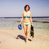 Na zdjęciu widać osobę w stroju kąpielowym, stojącą na piaszczystej plaży z maską do nurkowania i płetwami w rękach. W tle rozciąga się spokojne morze pod bezchmurnym niebem.