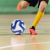 Na zdjęciu widać piłkę nożną z niebiesko-białym wzorem na hali sportowej oraz nogi osoby w żółtych getrach i różowych butach sportowych. Piłka jest w ruchu, a podłoga hali jest gładka i błyszcząca.
