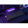 Na zdjęciu znajduje się klawiatura mechaniczna z podświetleniem RGB, umieszczona na ciemnym biurku. W tle widoczny jest kontroler do gier, mysz komputerowa oraz urządzenie z ekranem wyświetlającym kolorowe wzory.