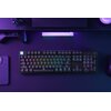 Na zdjęciu znajduje się klawiatura mechaniczna z podświetleniem RGB, umieszczona na ciemnym biurku, otoczona akcesoriami komputerowymi. Klawisze są podświetlone w różnych kolorach, a w tle widoczne są elementy oświetlenia LED.