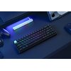 Na zdjęciu znajduje się klawiatura mechaniczna z podświetleniem RGB, umieszczona na ciemnym biurku. W tle widoczne są elementy takie jak kontroler do gier, słuchawki, myszka oraz tablet, a także podłużna lampka LED emitująca kolorowe światło.