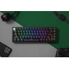 Na zdjęciu znajduje się klawiatura mechaniczna z podświetleniem RGB, umieszczona na dwukolorowym tle w odcieniach zieleni i szarości. Wokół klawiatury widoczne są słuchawki, smartfon, zegarek i kontroler do gier.