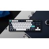 Na zdjęciu znajduje się klawiatura mechaniczna w kolorach czarno-biało-niebieskim z klawiszami oznaczonymi m.in. jako "esc", "tab", "shift", "enter", "backspace", "home", "end", "control", "option", "fn". W tle widoczne są okulary, obiektyw aparatu oraz g