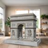 Na zdjęciu znajduje się model 3D Łuku Triumfalnego z puzzli, umieszczony na podstawce z napisem "ARC DE TRIOMPHE DE L'ÉTOILE". W tle widoczny jest jasny pokój z dużym oknem, rośliną i półkami.