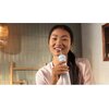 Osoba trzyma w ręku irygator PHILIPS Sonicare Compact Flosser 1000 HX3333/24 w kolorze niebieskim, używając go do czyszczenia zębów. W tle widoczne są kafelki i drewniany element dekoracyjny.